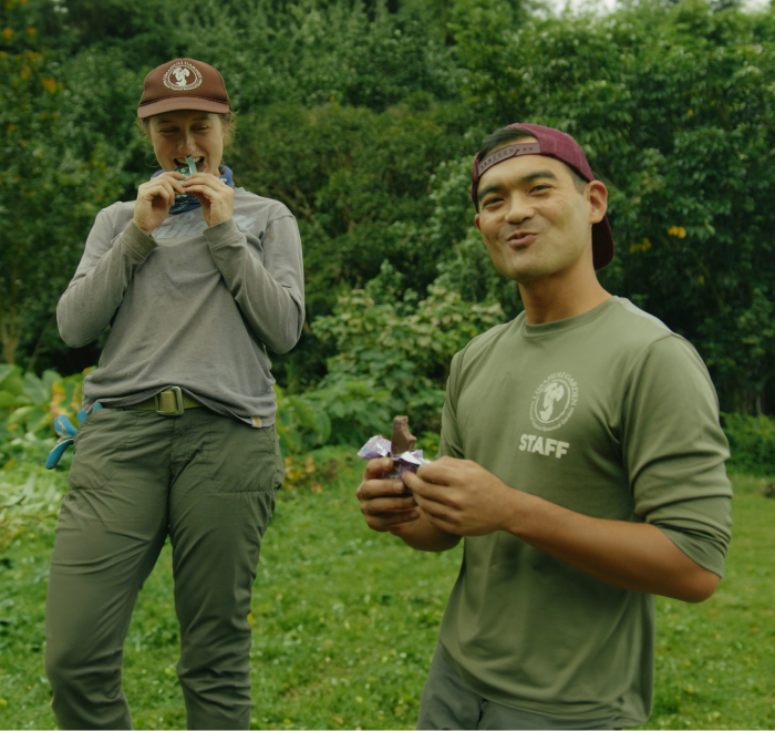 Two people enjoying snacks outdoors, standing on grass with greenery in the background.